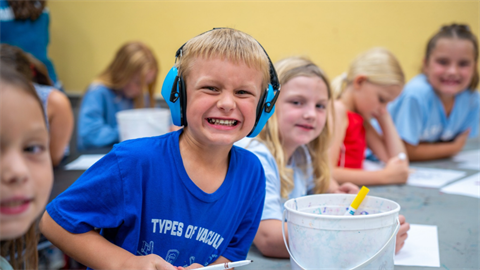 A smiling child wearing blue headphones participates in a craft activity with other children at camp.