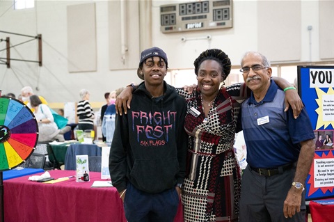 Two gentlemen with a woman in the middle smiling and posing in front of an exhibit table